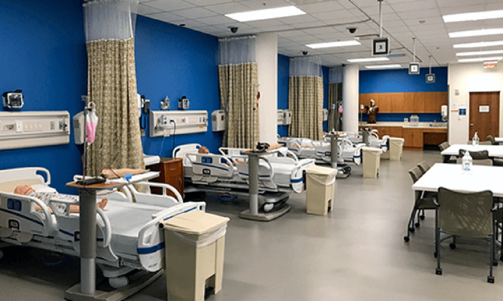 Four hospital beds set up in a row in a simulated hospital. Curtains are drawn back beside the beds and various medical equipment is around them. Conference tables and chairs are set up on the other side of the room.