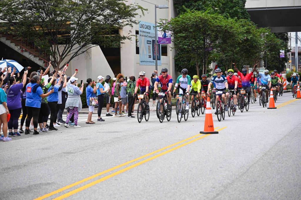 In this photo, you see people riding bicycles in the street during the PaceDay 2025 event to raise money for the Georgia Cancer Center. There are also people in this photo standing along the sidewalk cheering on the cyclists.