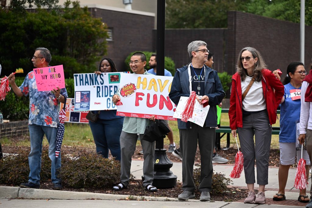 In this photo, you see men and women standing along a sidewalk holding signs to show support for cyclists participating in the PaceDay 2025 fundraising event.