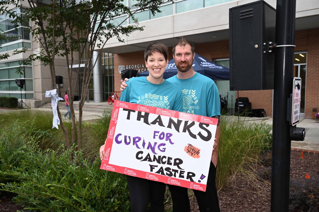 In this photo, you see two people holding a sign that reads "Thanks for curing cancer faster."
