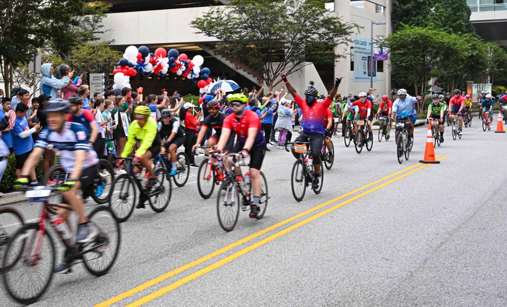 In this photo, you see people riding bicycles in the street during the PaceDay 2025 event to raise money for the Georgia Cancer Center. There are also people in this photo standing along the sidewalk cheering on the cyclists.