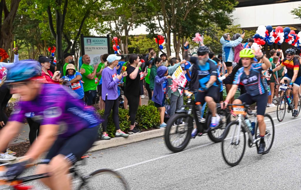 In this photo, you see people riding bicycles in the street during the PaceDay 2025 event to raise money for the Georgia Cancer Center. There are also people in this photo standing along the sidewalk cheering on the cyclists.