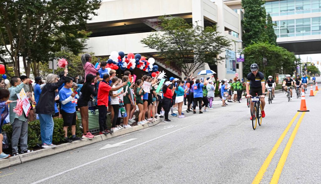 In this photo, you see people standing on the sidewalk to cheer on cyclists taking part in the PaceDay 2025 fundraising event for cancer research projects at the Georgia Cancer Center.