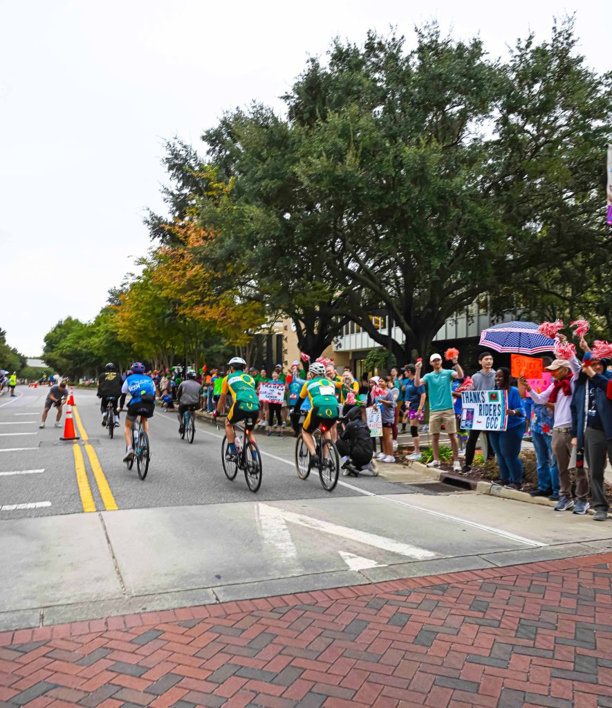 In this photo, you see people riding bicycles in the street during the PaceDay 2025 event to raise money for the Georgia Cancer Center. There are also people in this photo standing along the sidewalk cheering on the cyclists.