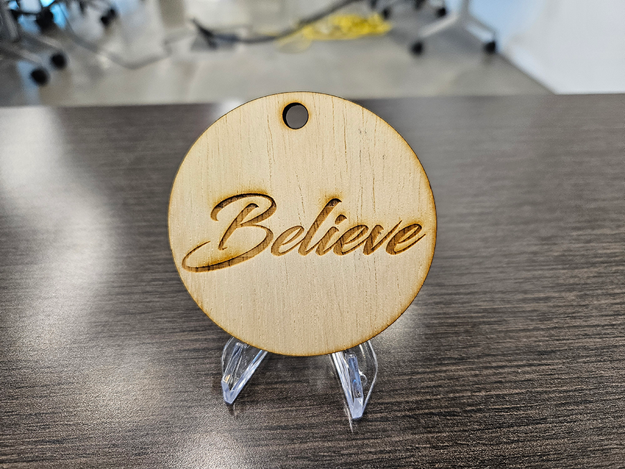 A holiday ornament made out of wood. The word "Believe" has been burned into the circular ornament.