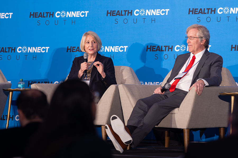 A woman and a man sit in chairs on a stage in front of a large group of people gathered for a health care conference. The woman is speaking into a microphone while the man sits and listens.