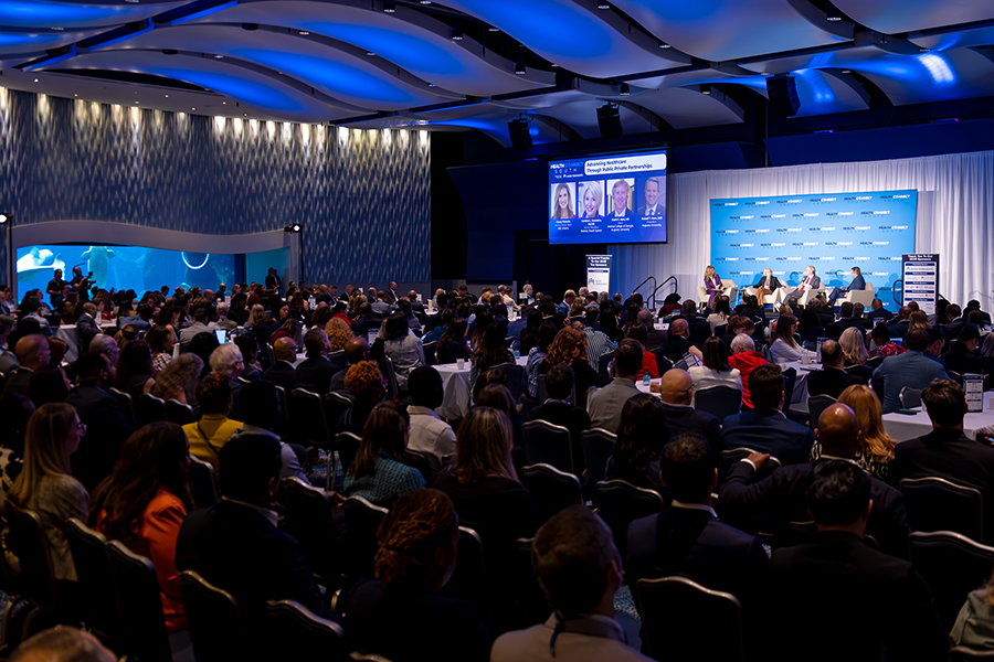 A group of people gather in a large conference room for a health care conference. One wall of the space features a window into an aqarium where beluga whales are swimming.
