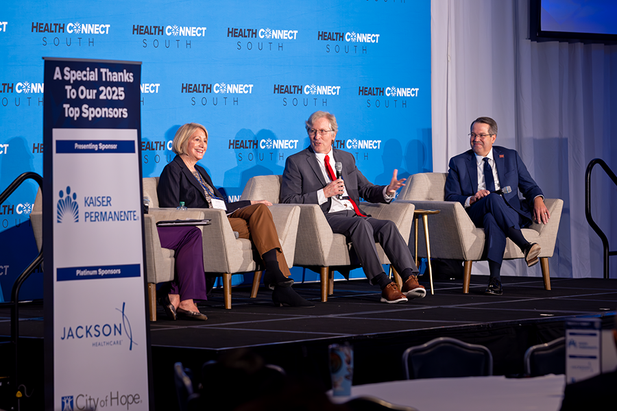Two men and two women sit in chairs on a stage in front of a large group of people gathered for a health care conference. One of the men is speaking into a microphone while the other three individuals on the stage sit and listen.