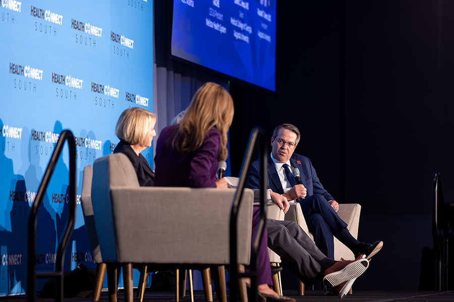 Two men and two women sit in chairs on a stage in front of a large group of people gathered for a health care conference. One of the men is speaking into a microphone while the other three individuals on the stage sit and listen.