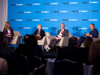 Two men and two women sit in chairs on a stage in front of a large group of people gathered for a health care conference. One of the men is speaking into a microphone while the other three individuals on the stage sit and listen.