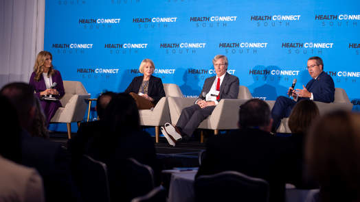 Two men and two women sit in chairs on a stage in front of a large group of people gathered for a health care conference. One of the men is speaking into a microphone while the other three individuals on the stage sit and listen.