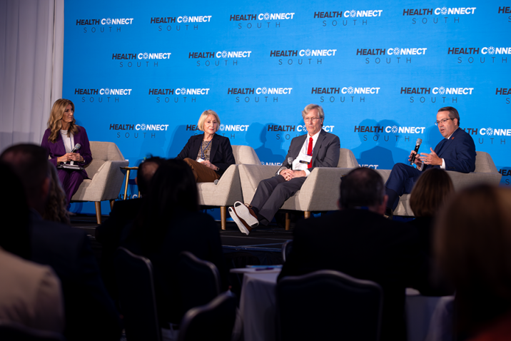 Two men and two women sit in chairs on a stage in front of a large group of people gathered for a health care conference. One of the men is speaking into a microphone while the other three individuals on the stage sit and listen.