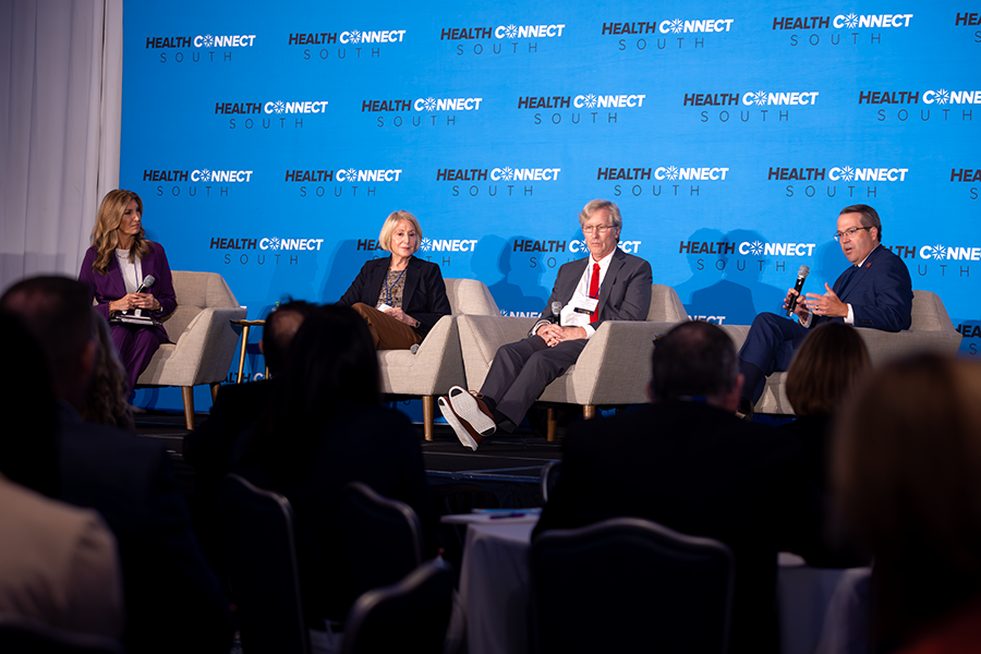 Two men and two women sit in chairs on a stage in front of a large group of people gathered for a health care conference. One of the men is speaking into a microphone while the other three individuals on the stage sit and listen.