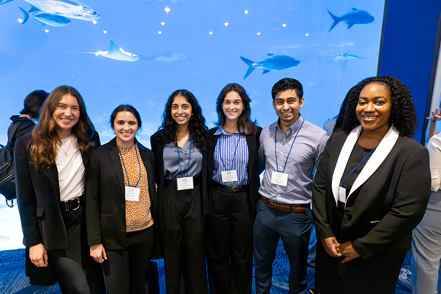 A group of four college-aged women, a college man, and their female professor stand in a large lobby of an aquarium with fish swimming in a large tank behind them.