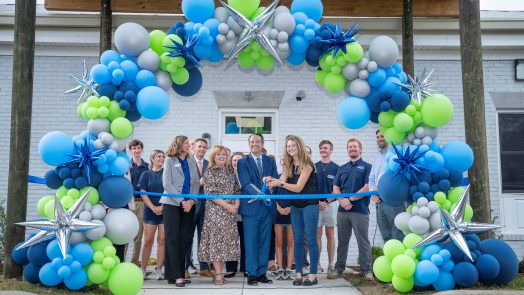 A group of men and women standing behind a large balloon arch as a man and woman prepare to cut the ribbon with scissors.