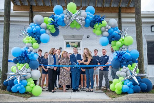 A group of men and women standing behind a large balloon arch as a man and woman prepare to cut the ribbon with scissors.