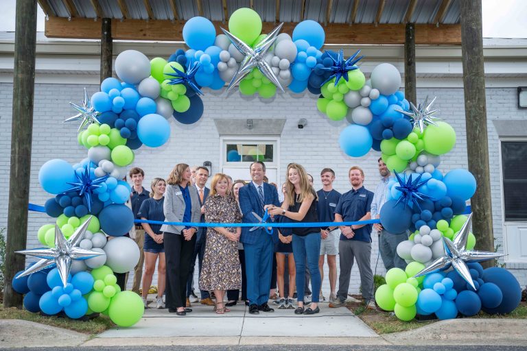 A group of men and women standing behind a large balloon arch as a man and woman prepare to cut the ribbon with scissors.