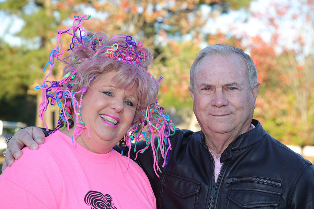 A man in a black leather coat puts his arm around a woman wearing a bright pink shirt with neon colored beads and strings throughout her pink hair.