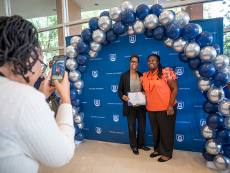 Two women stand under a balloon arch during a celebration while another woman takes their photo.