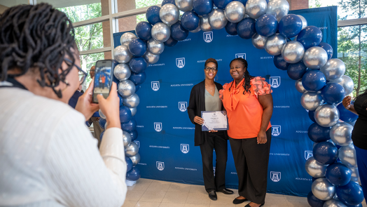 Two women stand under a balloon arch during a celebration while another woman takes their photo.