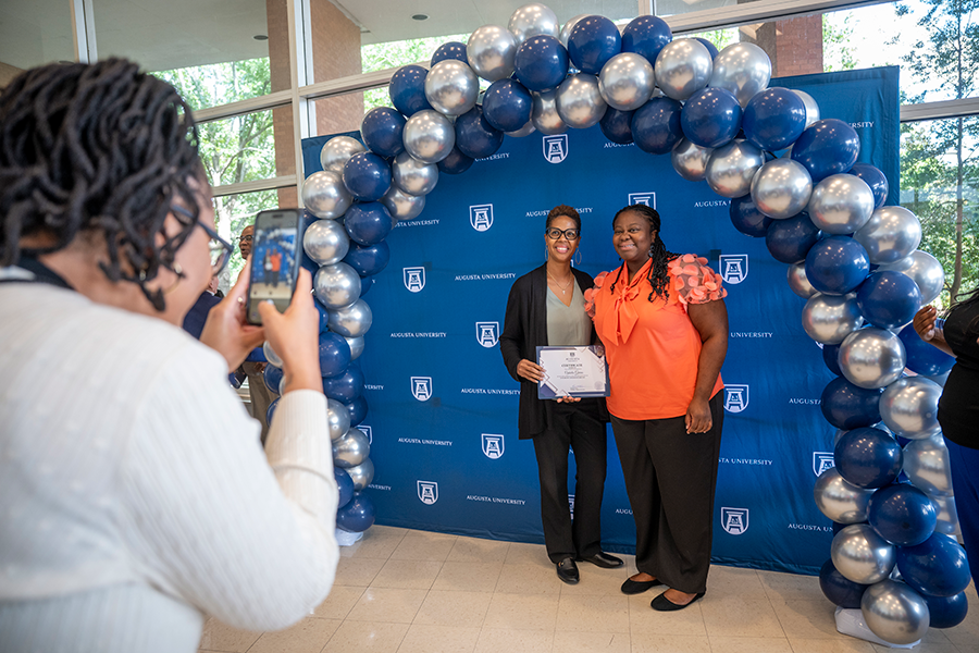 Two women stand under a balloon arch during a celebration while another woman takes their photo.