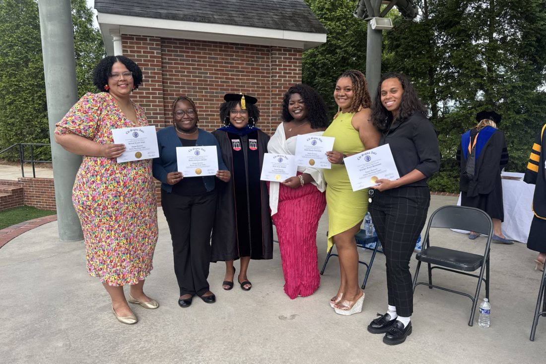 A group of five women stand on an outdoor stage during a ceremony. All five are holding up paper certificates of completion.