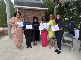 A group of five women stand on an outdoor stage during a ceremony. All five are holding up paper certificates of completion.