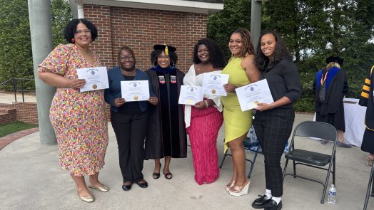 A group of five women stand on an outdoor stage during a ceremony. All five are holding up paper certificates of completion.