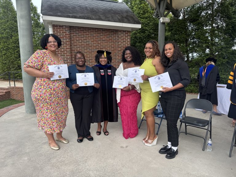 A group of five women stand on an outdoor stage during a ceremony. All five are holding up paper certificates of completion.