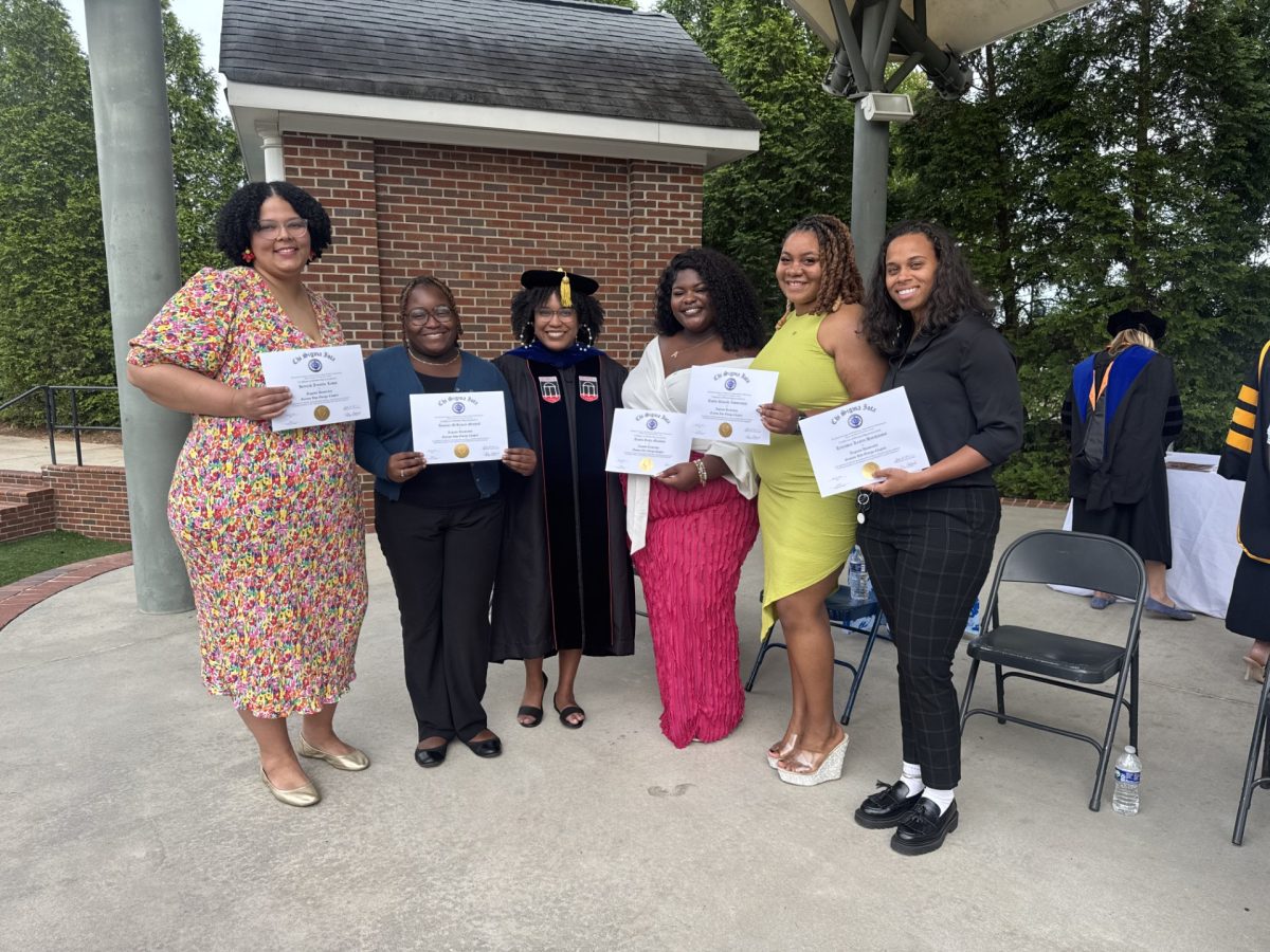 A group of five women stand on an outdoor stage during a ceremony. All five are holding up paper certificates of completion.