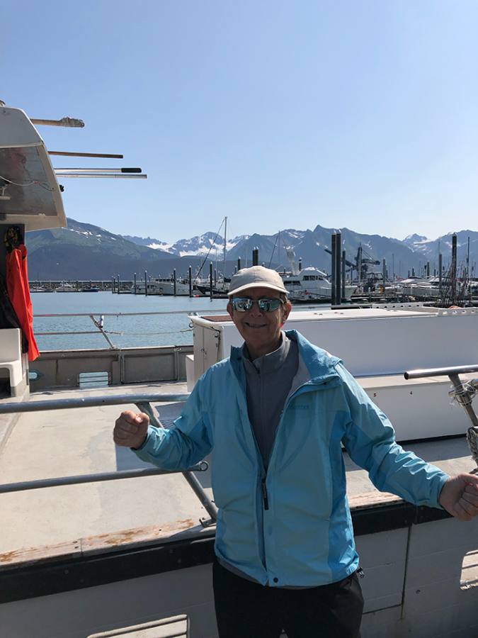 A woman wearing a jacket and hat poses in front of a dock of boats and a snowy mountain range.