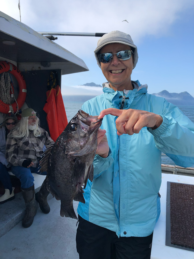 A woman wearing a jacket and hat holds up a rock cod she caught while fishing in Alaska.