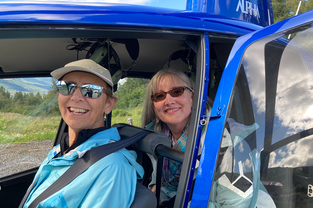 Two women sit in a helicopter and smile at the camera.