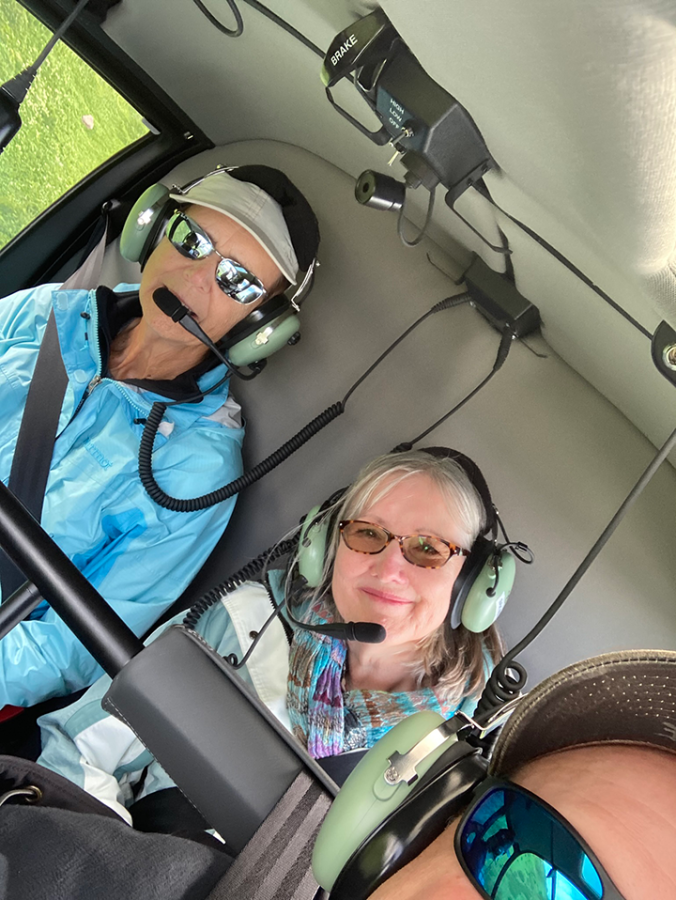 Two women sit in the back of a helicopter wearing headsets.