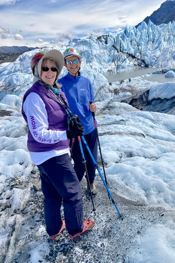Two women wearing cold weather gear and holding walking sticks pose for a photo on an icy glacier in Alaska.