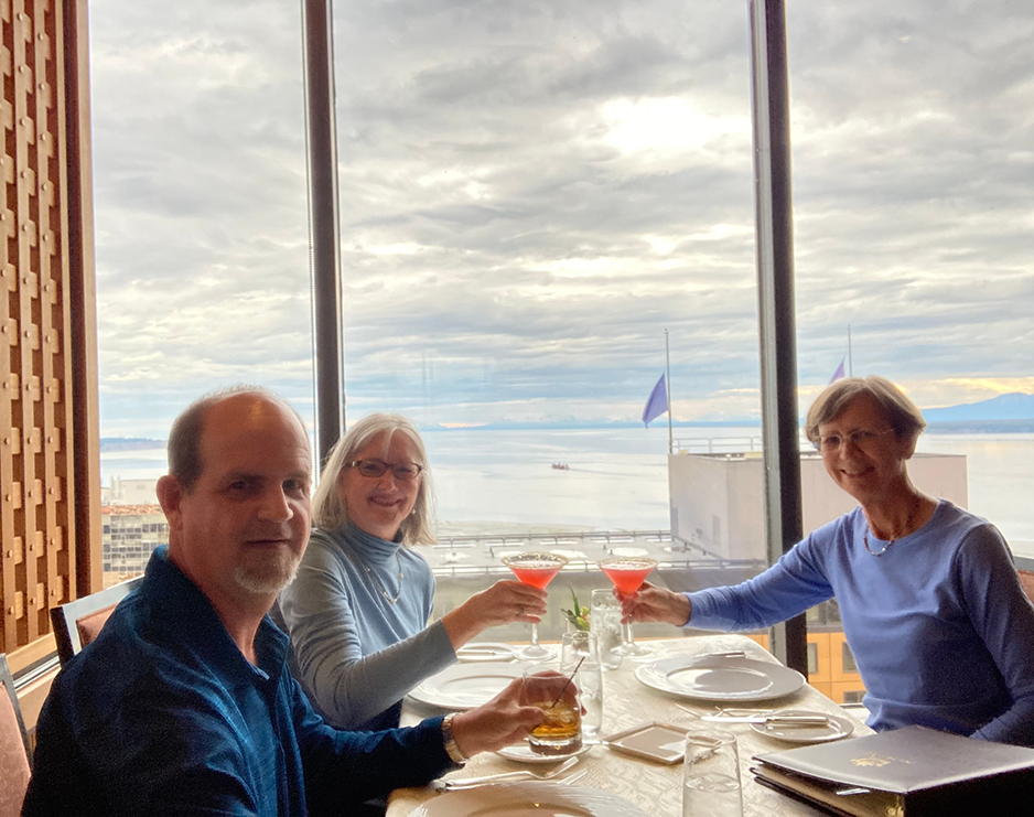 Two women and a man drink cocktails and sit at a table in front of a view of an ocean and mountain range.