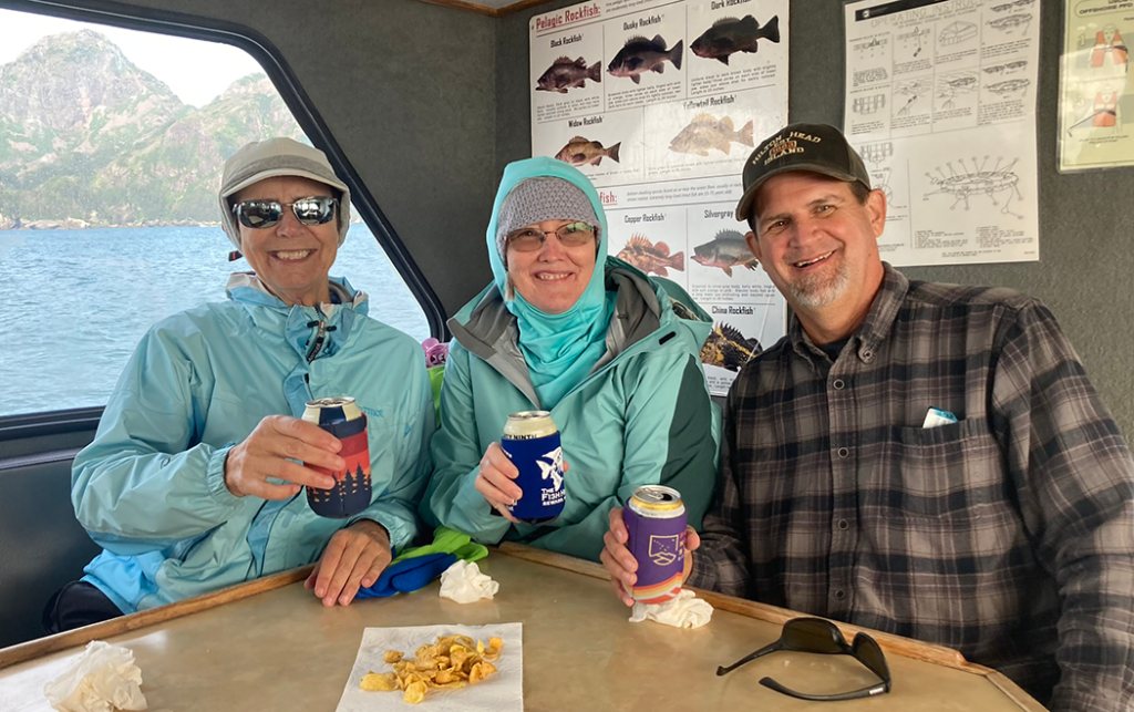 Two women and a man wearing jackets sit at a table on a boat and hold up cans of drinks for a toast.