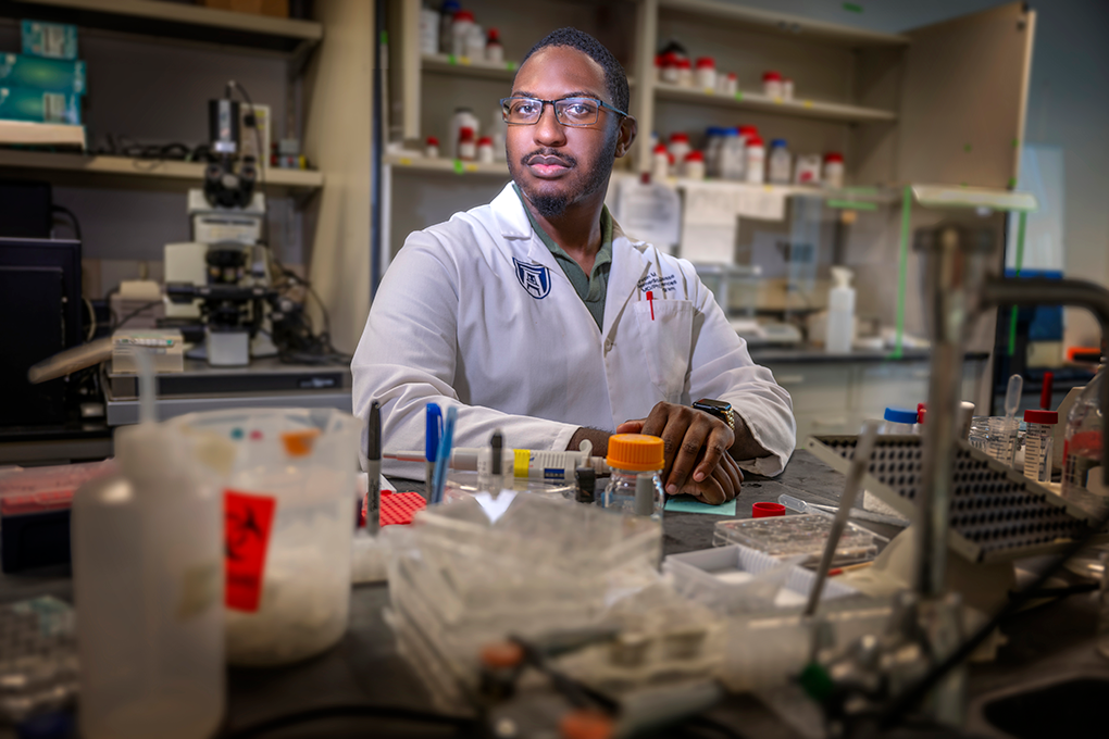 A man wearing glasses and a lab coat sits at a table in a laboratory. There are beakers and other research materials on the table.