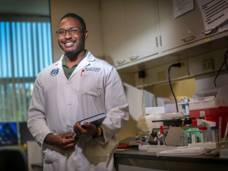 A man wearing glasses and a lab coat holds a book and stands in front of a table in a laboratory.