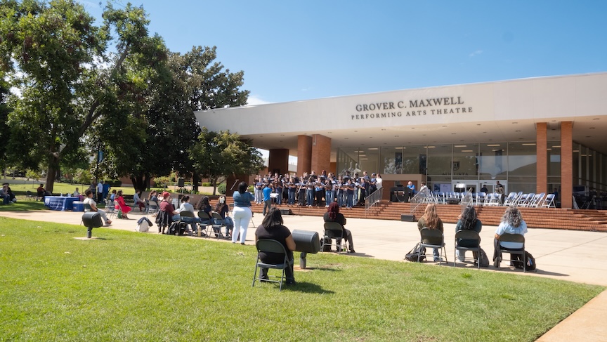 A group of people sit outside in chairs and enjoy a musical concert. They are outside a performing arts building with "Grover C. Maxwell Performing Arts Theatre" in large letters on the facade of the building.