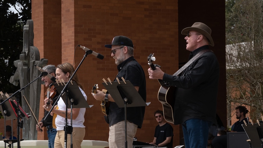 Three men and a woman play instruments as part of a band. One man and the woman are playing guitars, one man is playing a mandolin and the last man is holding a violin.