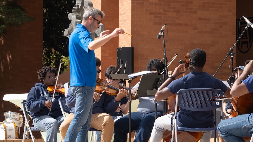 A male musical conductor directs a band during an outdoor performance.