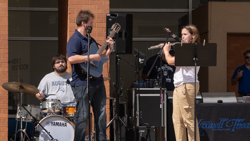 Three college students, two men and one woman, play musical instruments during an outdoor performance. One man is playing the drums, one is on a guitar, and the woman is playing the flute.