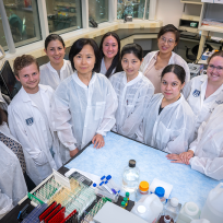 A group of researchers wearing lab coats gather around a lab table. On the table, there are vials and lab gear.