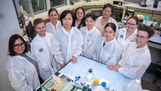 A group of researchers wearing lab coats gather around a lab table. On the table, there are vials and lab gear.
