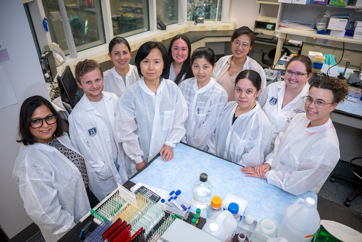 A group of researchers wearing lab coats gather around a lab table. On the table, there are vials and lab gear.