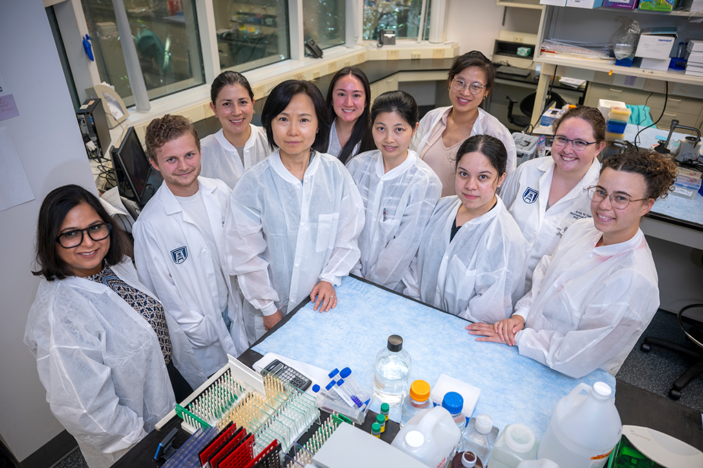 A group of researchers wearing lab coats gather around a lab table. On the table, there are vials and lab gear.