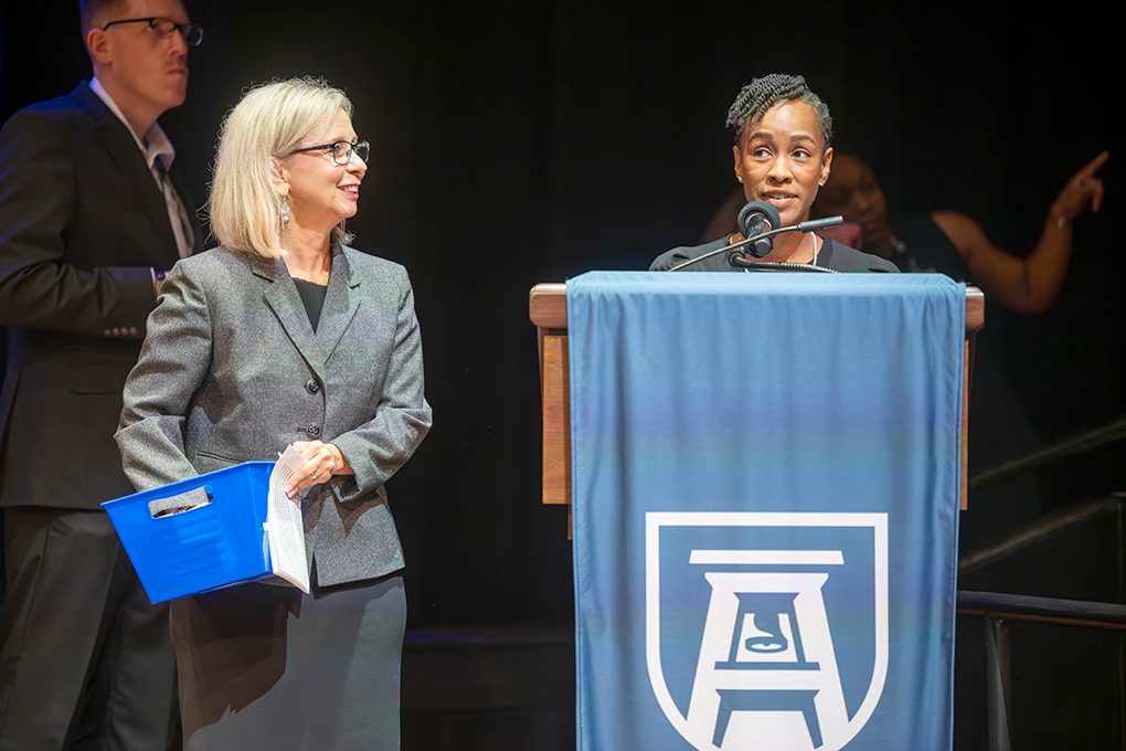 A woman with her hand pulling something out of a box and a woman speaking into a microphone behind a lectern.