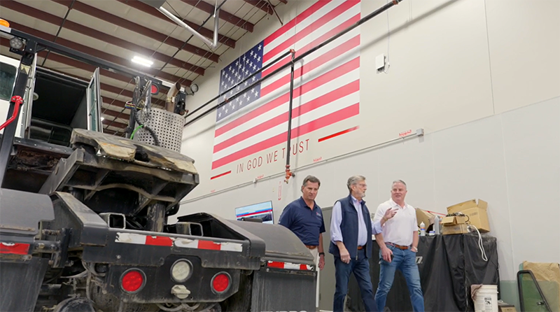 Three men walk through a shop where tractor trailer trucks are being serviced.
