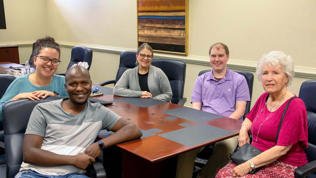An older woman sits at a conference table with two college-aged women and two college-aged men.